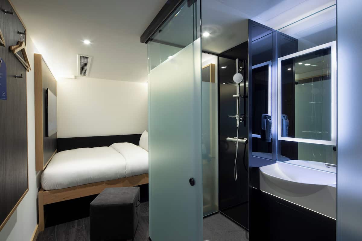 A modern hotel room with a bed, a glass partition, a shower area, and a bathroom with a sink and mirror.