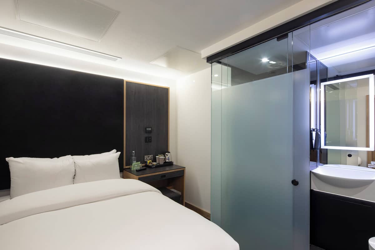 A modern hotel room with a bed, bedside table, and a glass-enclosed bathroom with a sink and mirror.