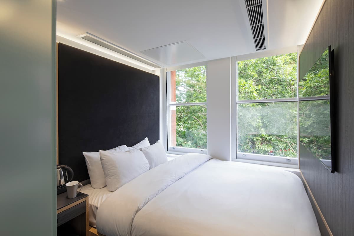 A modern bedroom with a large bed, a nightstand, a cup, and a window with a view of greenery.
