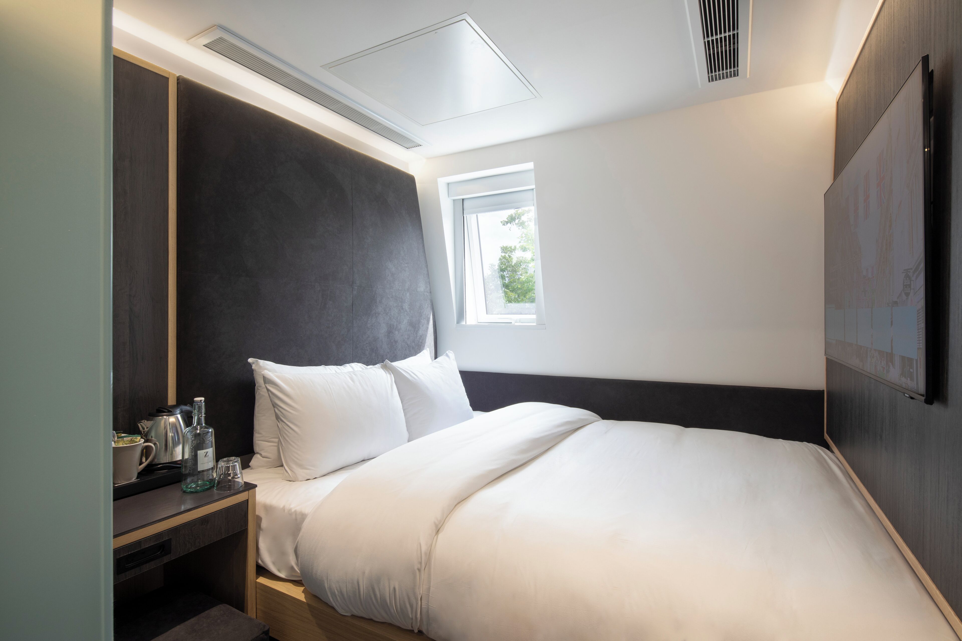 Ein modernes Hotelzimmer mit einem großen Bett, einem Nachttisch mit Wasserkocher und Gläsern, einem Fenster mit Blick auf Grünflächen und einem an der Wand befestigten Fernseher.
