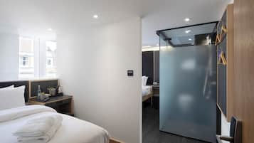 Ein modernes Hotelzimmer mit einem ordentlich bezogenen Bett, einem kleinen Nachttisch mit einer Lampe, einem Kleiderschrank mit Spiegel und einer Schiebetür zum Badezimmer.