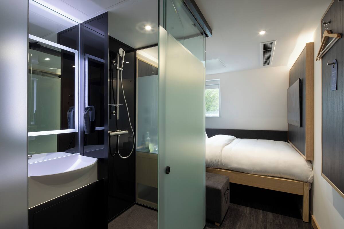 A modern hotel room with a glass shower enclosure, a built-in sink, a bed with a wooden frame, and a large mirror.