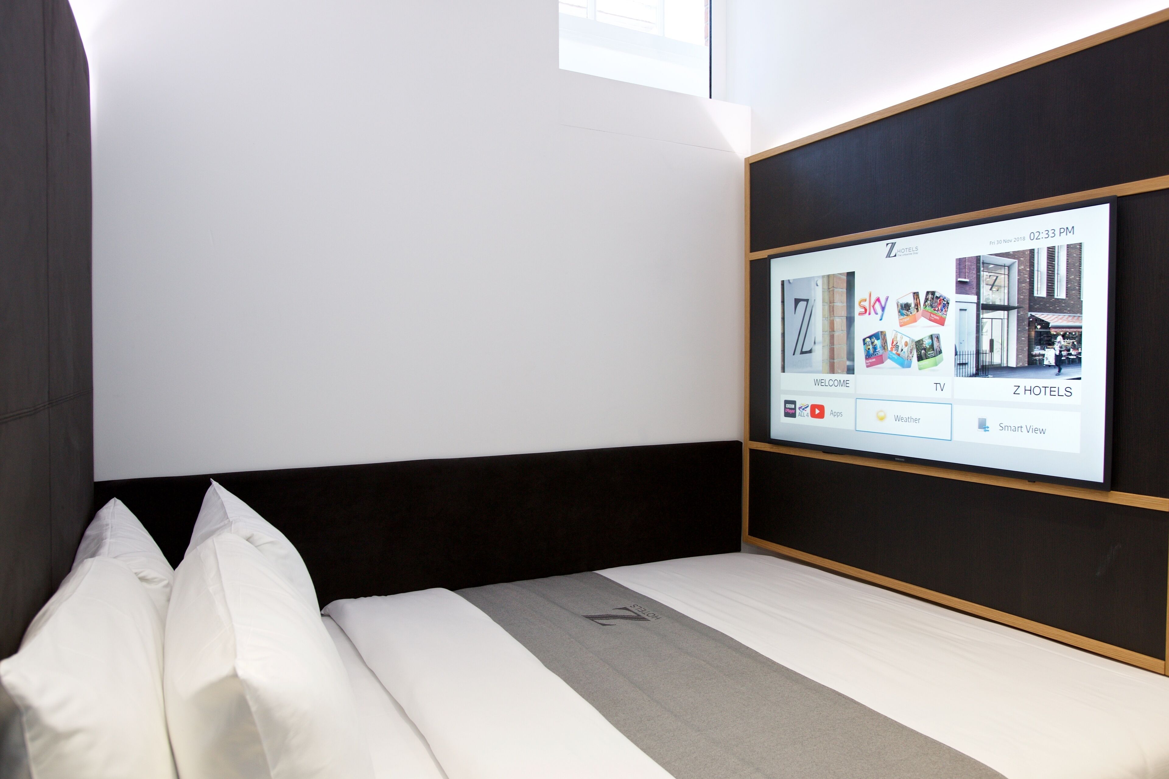 A modern hotel room with a large TV screen displaying the hotel's welcome page, a neatly made bed with white linens, and a dark headboard.