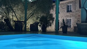 Una piscina con zona techada, edificio de piedra y plantas en macetas.