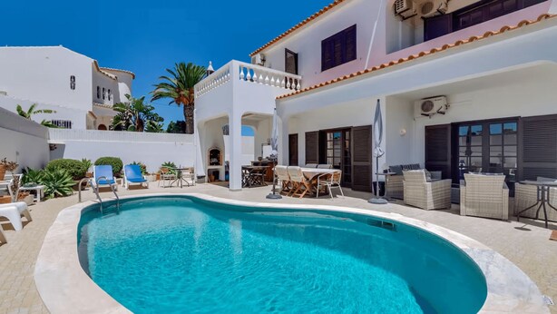 Una piscina amb gandules i taules, envoltada d'edificis blancs amb balcons i palmeres.