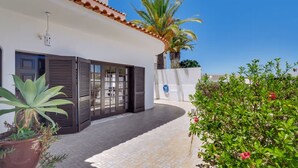 Una casa moderna amb un pati enrajolat, una gran porta corredissa de vidre i un jardí amb plantes i una palmera.