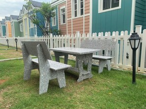 Un banco de piedra y una mesa ubicados en un área verde, con una cerca de piquetes blancos y casas coloridas al fondo.