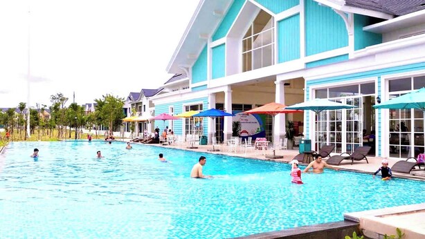 A swimming pool area with lounge chairs and umbrellas, a building with large windows, and a clear sky. - Deluxe 3 Br Happy House - Novaworld PT (Tien Thanh)