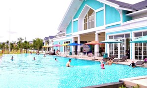 A swimming pool area with lounge chairs and umbrellas, a building with large windows, and a clear sky. - Deluxe 3 Br Happy House - Novaworld PT (Tien Thanh)
