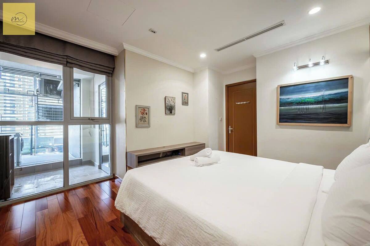 A hotel room with a bed, a painting on the wall, and a window with a view of the outside.
