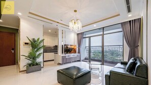 A modern living room with a sofa, ottoman, and a large potted plant. There is a dining area with a TV and a balcony with a view. - Landmark 81 Magical Tower Apartment (Ho Chi Minh City)