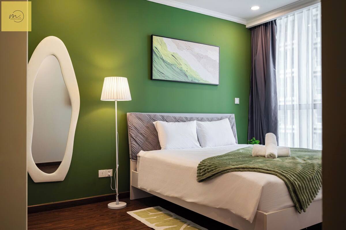 A modern hotel room with a green wall, a large bed with white bedding, a bedside lamp, and a painting above the bed.
