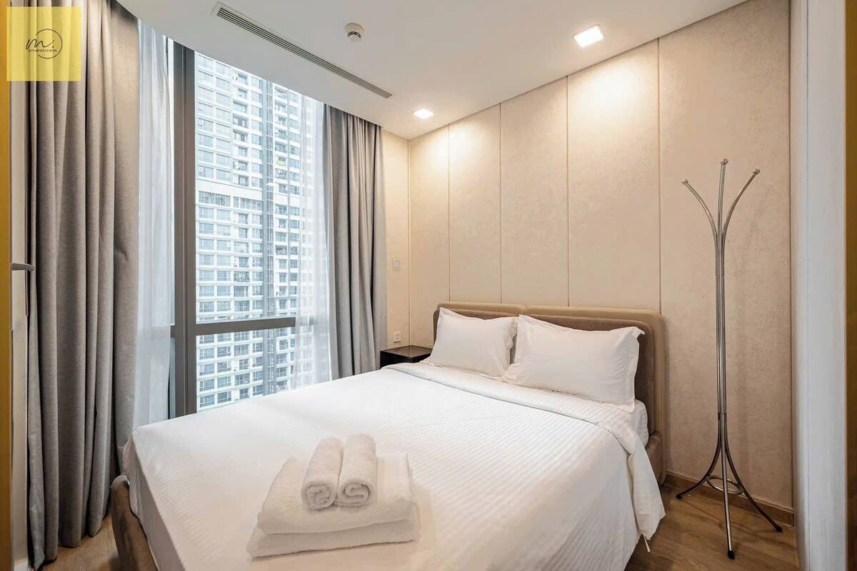 A hotel room with a large bed, a standing lamp, and a view of a high-rise building through the window.