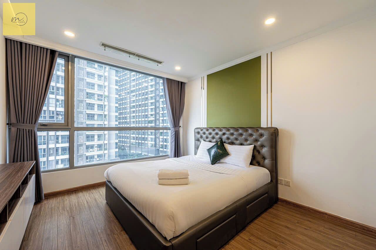 A modern hotel room with a large bed, a desk, and a view of a cityscape through large windows.