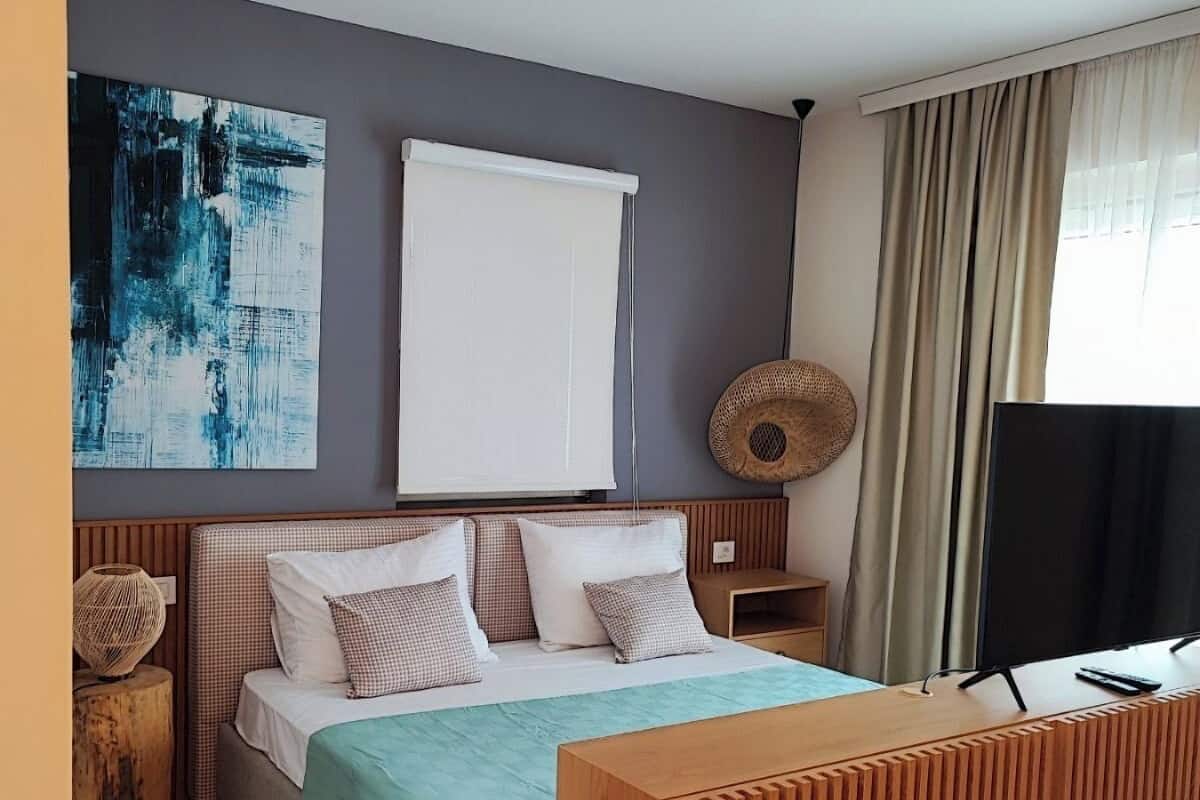 A modern hotel room with a bed, a wooden side table, a woven basket, a large abstract painting, a white ceiling, and a window with curtains.