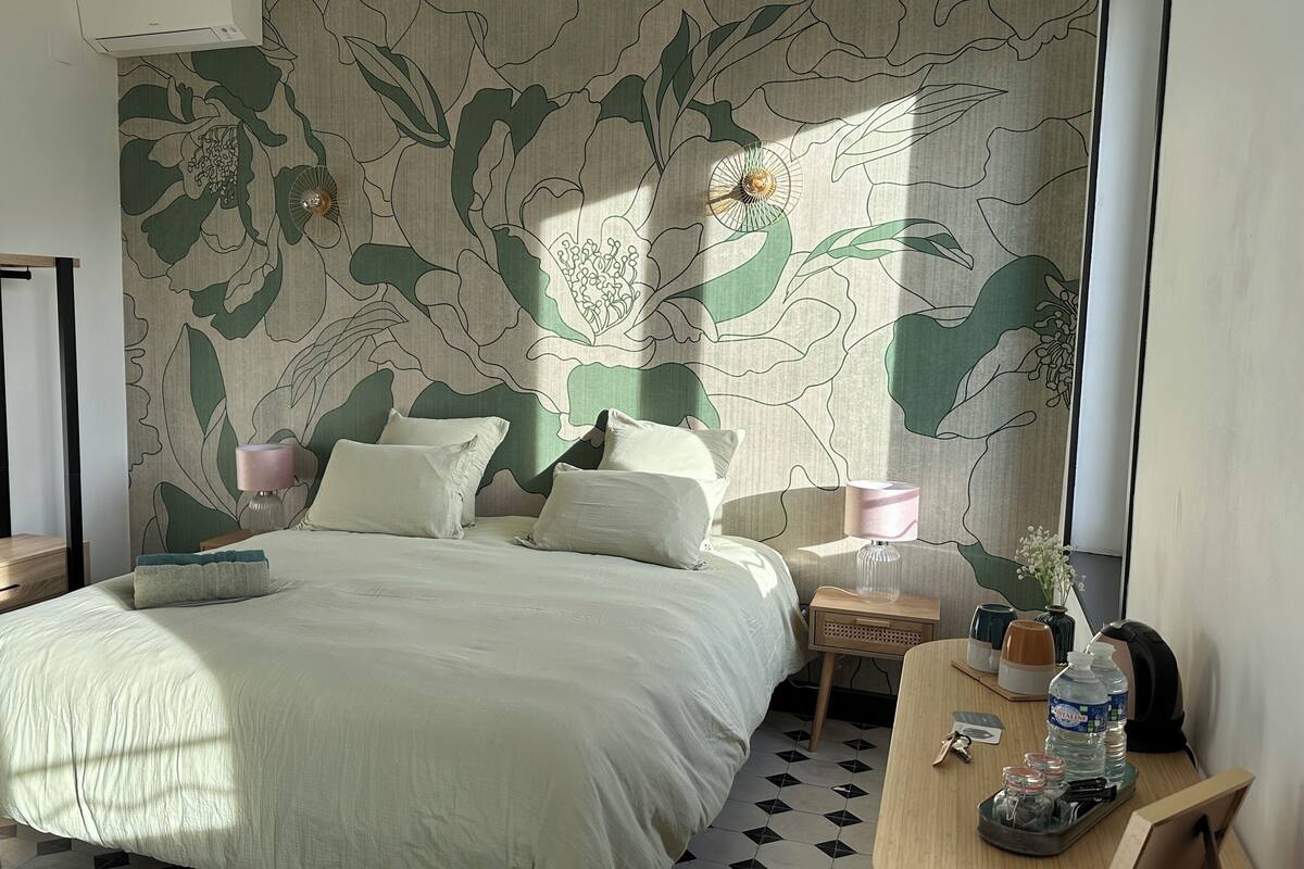 Un dormitorio con cama, mesitas de noche, una lámpara, una pequeña mesa con un florero y botellas, y un papel tapiz con diseño de hojas.
