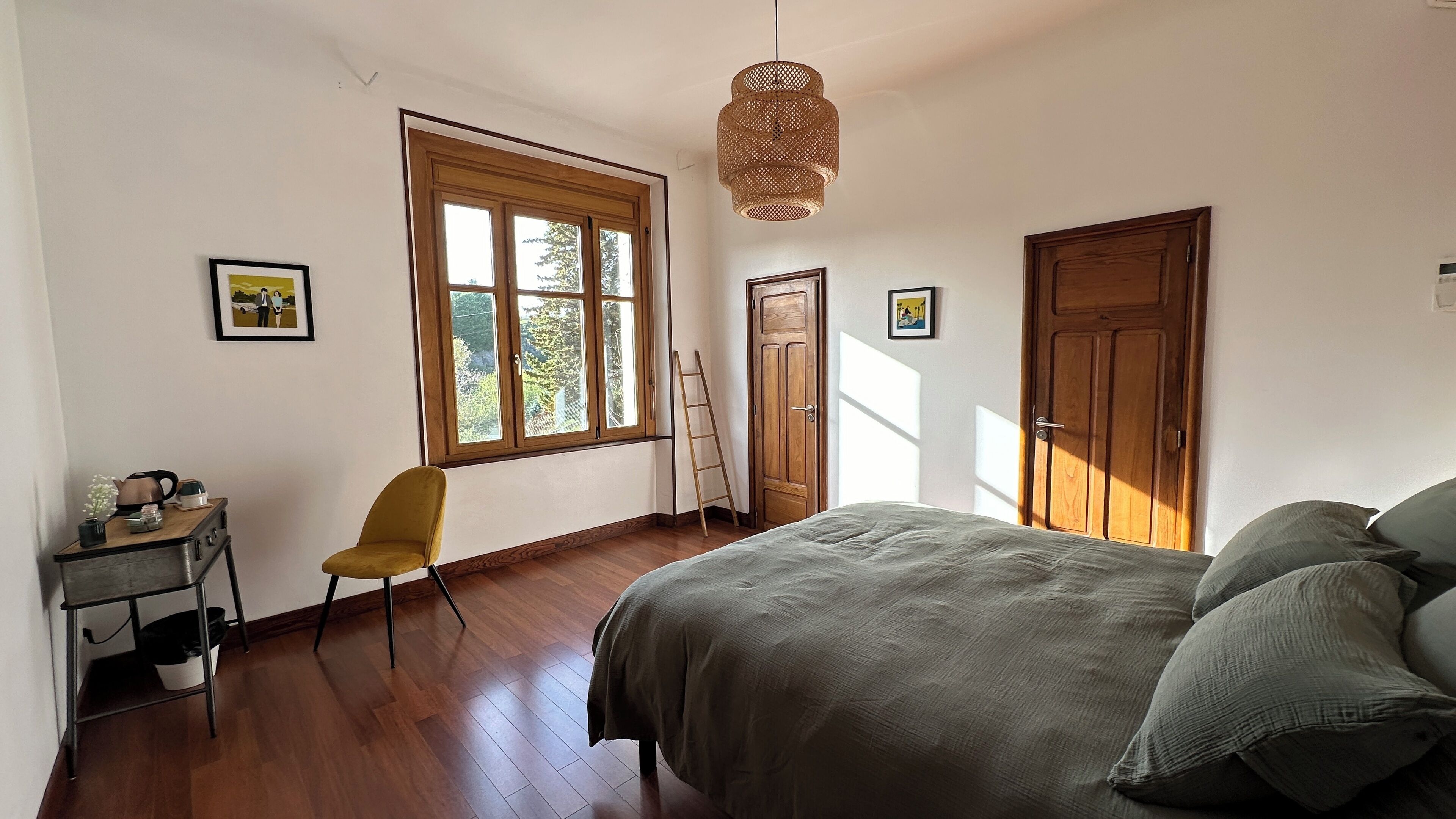 A bedroom with a bed, a chair, a desk with a kettle, a ladder, and a window with a view of trees.