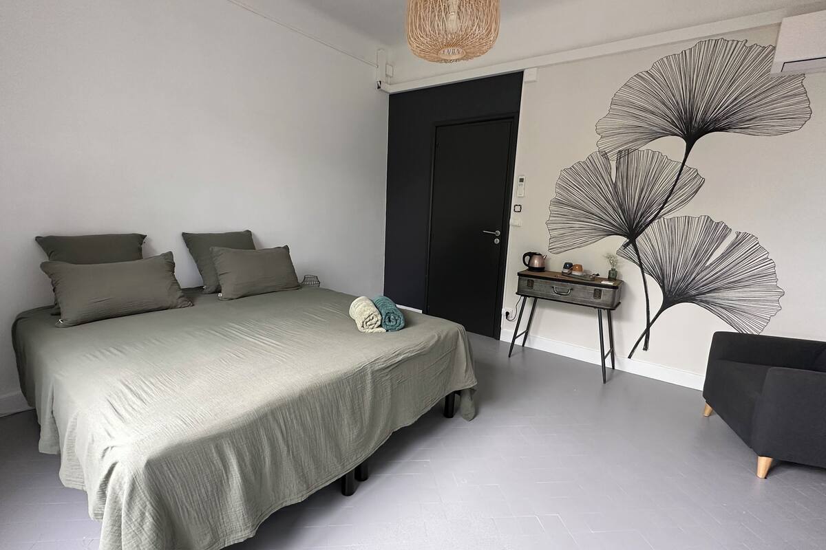 Un dormitorio con cama, almohadas, una mesita de noche, una silla y una obra de arte en la pared.