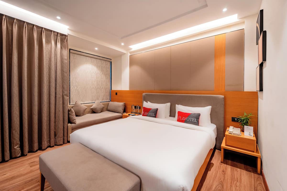 Una habitación de hotel moderna con una cama grande, un sofá, una mesita de noche de madera y un televisor montado en la pared.