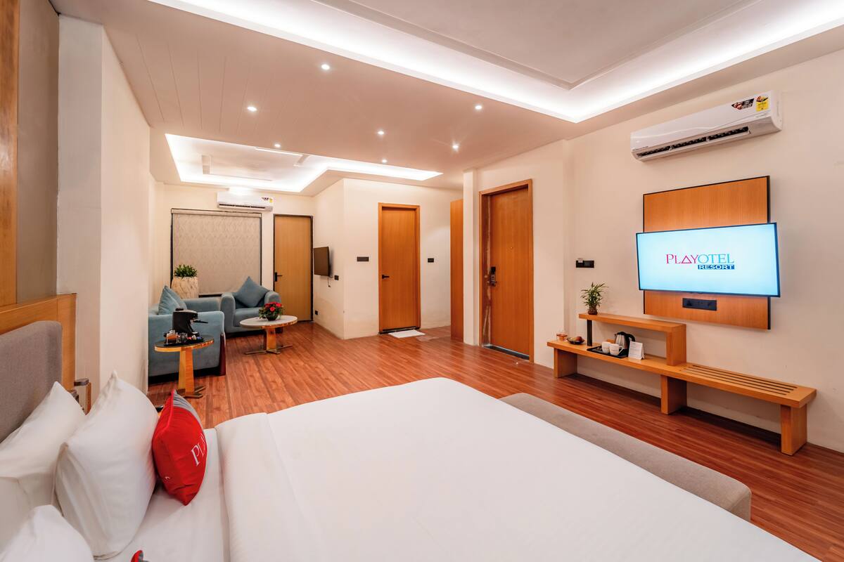 Una habitación de hotel moderna con una cama grande, un televisor, una zona de estar con sofá y mesa de centro, y un banco de madera.
