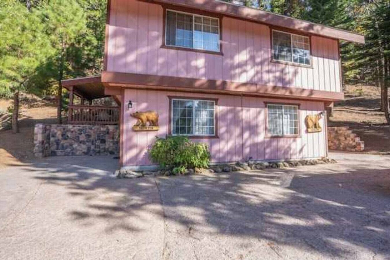 A two-story pink house with a stone foundation, a porch with a wooden railing, and two bear statues on the front.