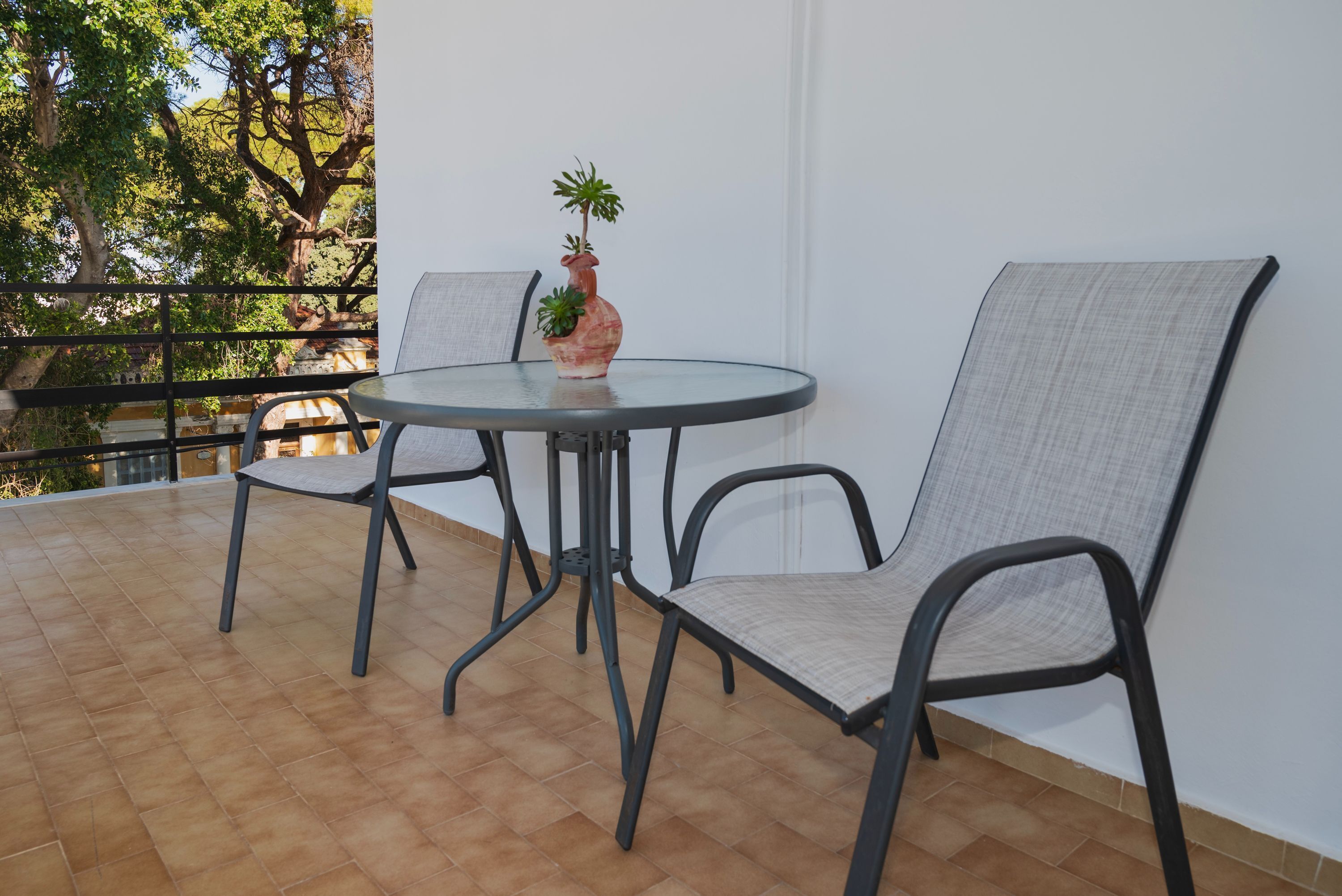 A round table with two chairs, a potted plant, and a view of trees and a balcony railing.