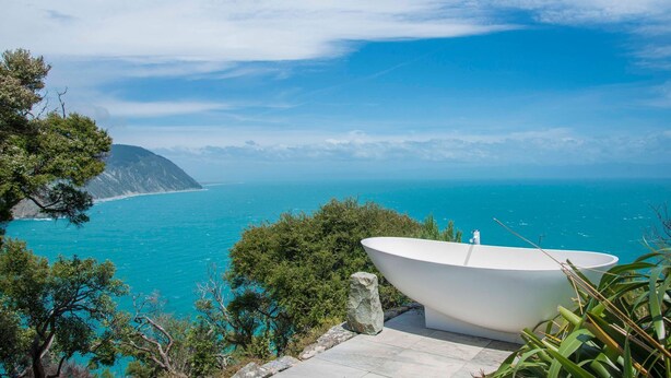 A white bathtub on a stone ledge overlooking a turquoise sea with a mountainous coastline in the background.