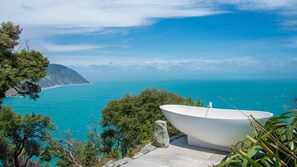 A white bathtub on a stone ledge overlooking a turquoise sea with a mountainous coastline in the background.