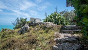 A stone pathway leading to a white bowl-shaped structure on a hillside with a view of the sea.