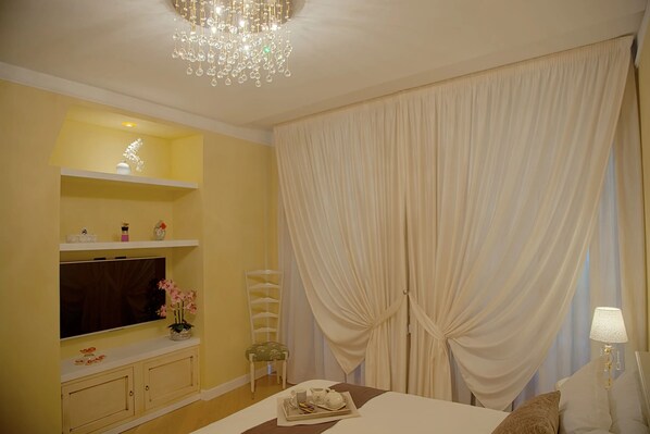 A hotel room with a large flat-screen TV, a chandelier, a bed with a tray of toiletries, and a large window with white curtains. - AMAZING FLORENCE APARMTENT (Firenze)