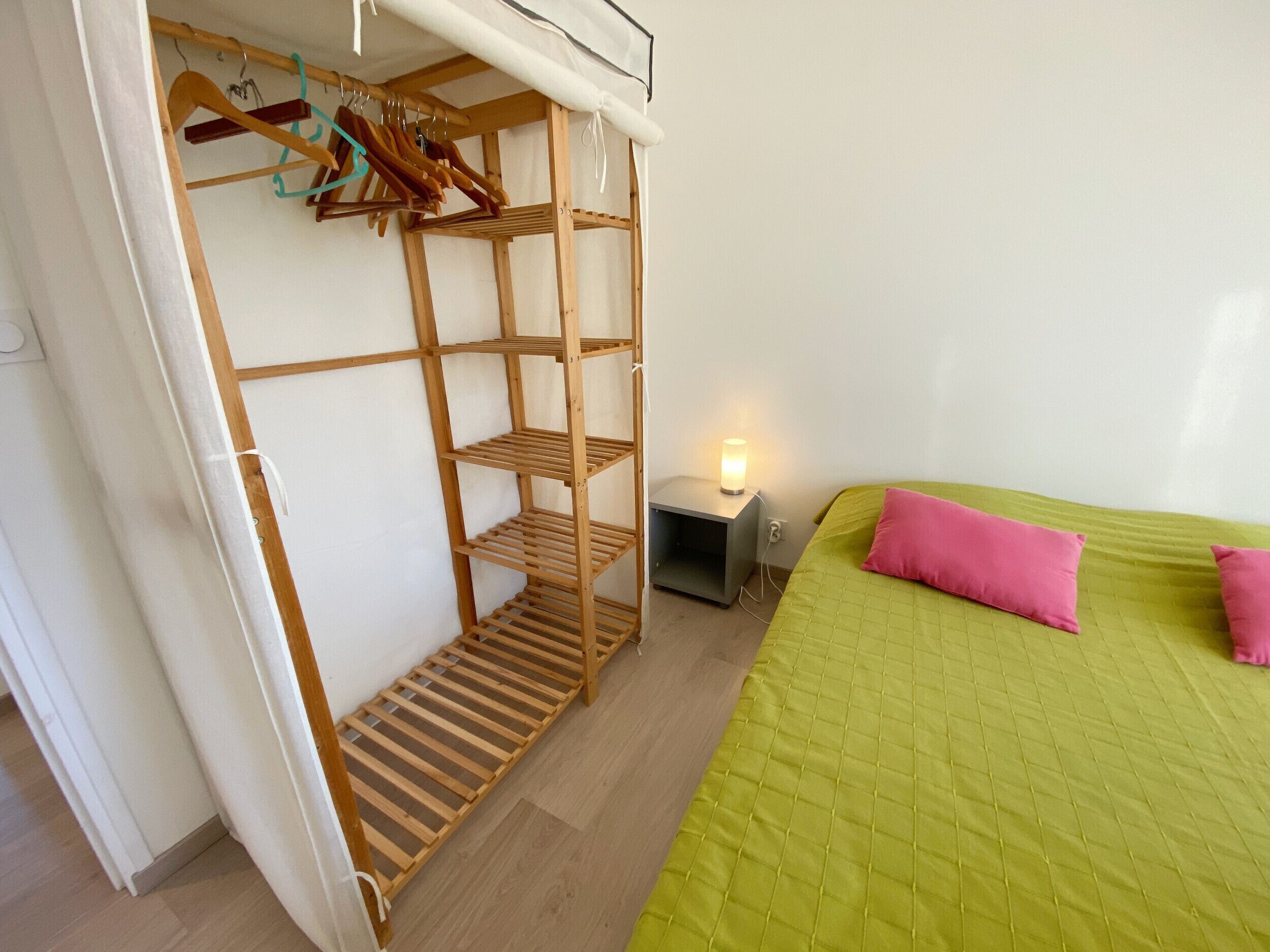 A bedroom with a wooden wardrobe, a bedside table with a lamp, and a green bed with pink pillows.