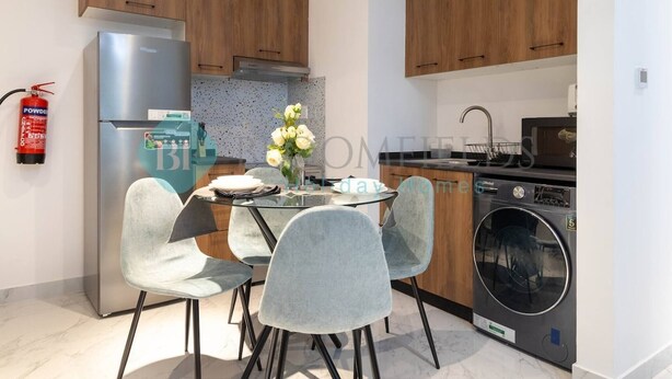 A modern kitchen with wooden cabinets, a stainless steel refrigerator, a washing machine, and a glass dining table with four chairs. - Bloomfields Stylish 1BR In Oasis Masdar City (Abu Dabi)