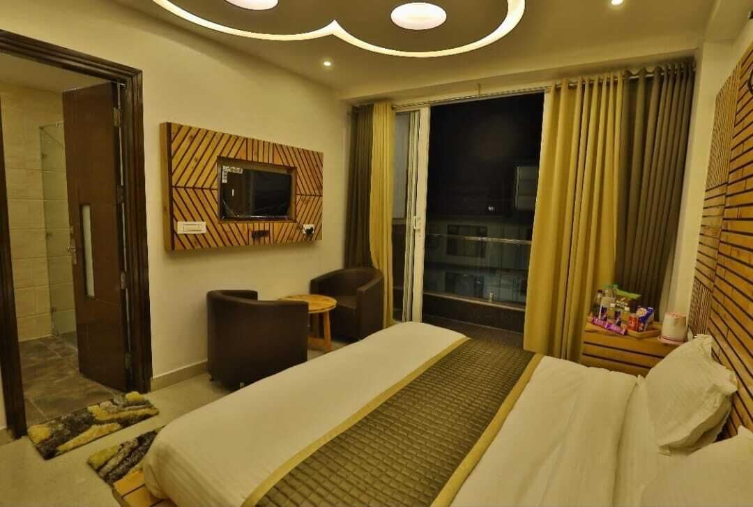 A hotel room with a bed, a desk, a chair, a TV mounted on the wall, and a window with curtains.