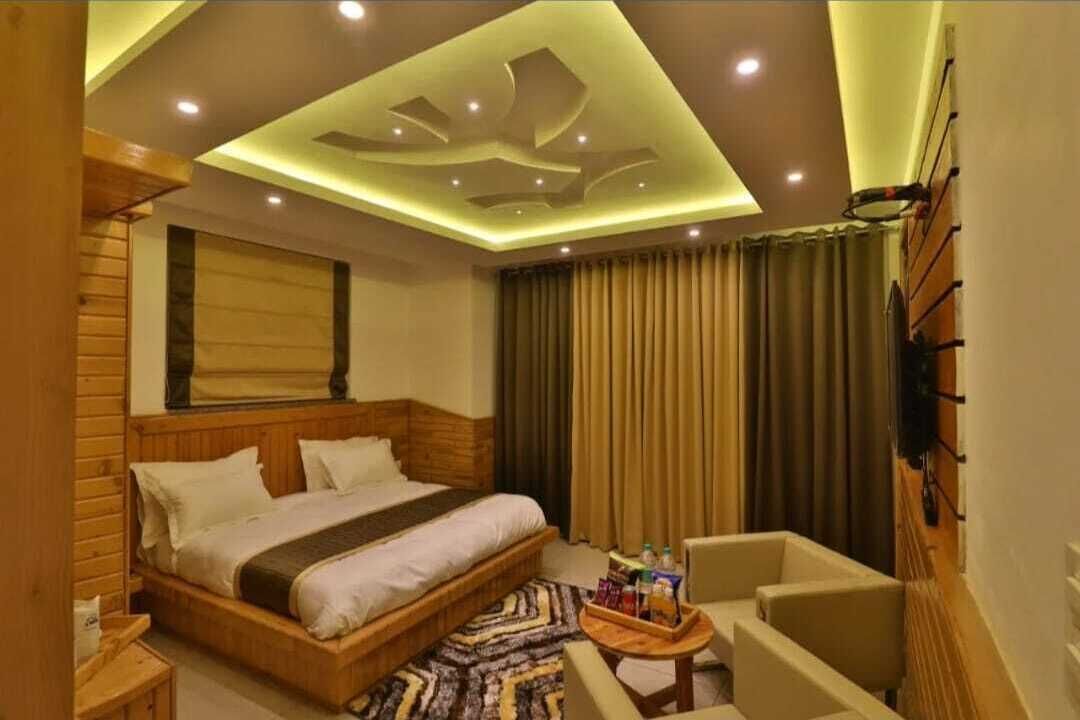 A hotel room with a wooden bed, a small seating area, a ceiling with recessed lighting, and a television mounted on the wall.