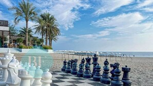 A chessboard set up on a beach with palm trees and a hotel in the background.