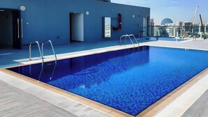 A rooftop swimming pool with a blue interior, surrounded by a modern building with a flat roof and a clear sky in the background.