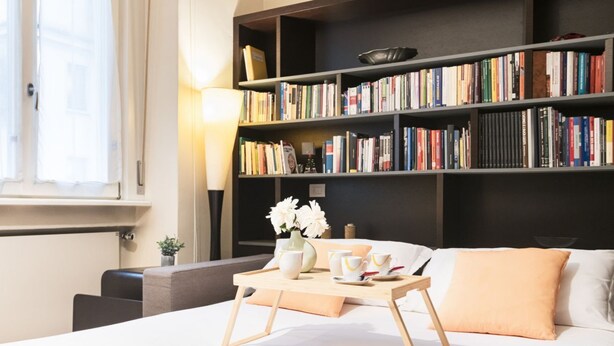 Een moderne hotelkamer met een groot bed, een nachtkastje met een ontbijtbakje, een boekenplank vol boeken en een raam met gordijnen.