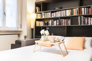 Una habitación de hotel moderna con una cama grande, una mesita de noche con una bandeja de desayuno, una estantería llena de libros y una ventana con cortinas.