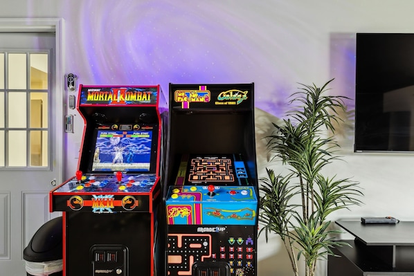 Dos máquinas de videojuegos arcade, una con el rótulo 'Mortal Kombat' y la otra 'Pac-Man', en una sala con una planta en maceta y un televisor en la pared.