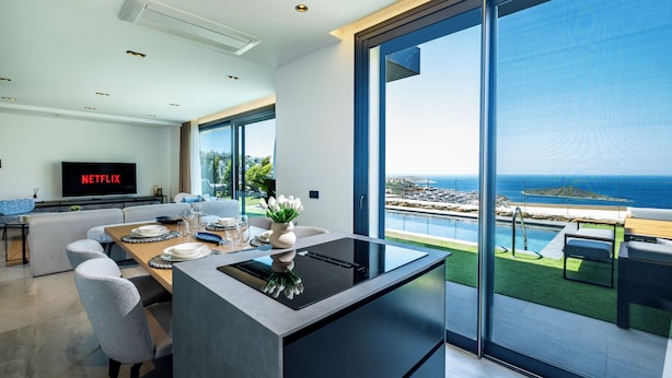 Una cocina moderna con una isla central, un área de comedor con una mesa dispuesta para cuatro personas y un amplio ventanal con vistas a una piscina y al océano.