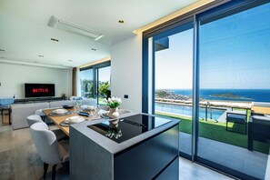 Une cuisine moderne dotée d’un îlot central, d’un coin repas avec une table pour quatre personnes et d’une grande fenêtre donnant sur une piscine et l’océan.