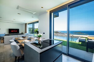 Uma cozinha moderna com uma ilha central, uma sala de jantar com mesa posta para quatro pessoas e uma grande janela com vista para uma piscina e oceano.