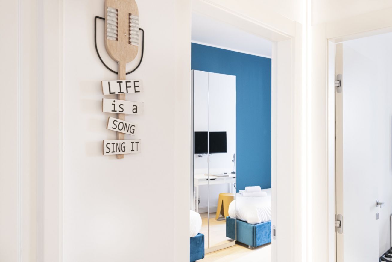 A modern hotel room with a blue accent wall, a wooden wall art piece with the phrase "LIFE is a SONG SING IT", a bed, a desk with a computer, and a mirror.