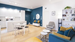 A modern living room with a blue sofa, a grey armchair, and a yellow ottoman. There is a dining area with a white table and chairs, and a kitchen area with white cabinets and a microwave. - Easylife: Elegant with Terrace near Centrale FS (Milano)