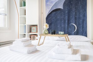 Cama arrumada com toalhas dobradas, mesa de cabeceira com um vaso e xícaras, e uma estante com livros e quadros.