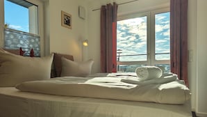 A hotel room with a bed, a window with curtains, a wall-mounted light, and a framed picture. - Marina-Penthouse 625 - Spacious vacation apartment on 1 level with harbor view. (Rechlin-Nord)