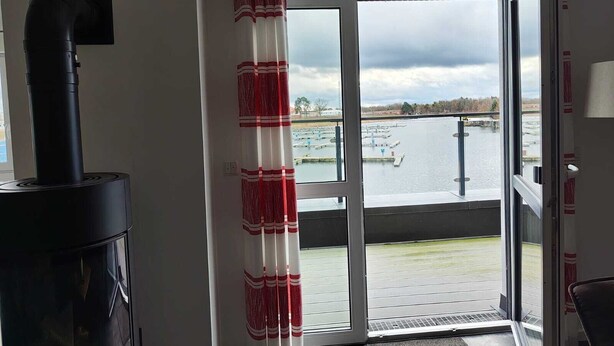 A room with a black wood stove, a large sliding glass door leading to a docked boat, and a view of a body of water. - Marina-Penthouse 625 - Spacious vacation apartment on 1 level with harbor view. (Rechlin-Nord)