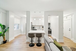 Moderni keittiö, jossa on valkoiset kaapit, keskellä sijaitseva saareke baarituoleineen sekä ruokailutila, jossa on harmaa sohva.