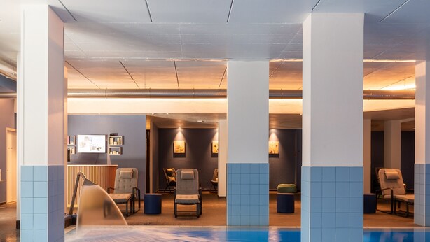 A modern indoor pool area with a blue pool, seating area with chairs, and a reception desk with a computer monitor.