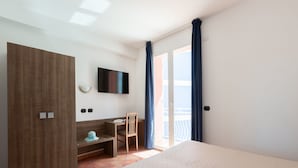 Une chambre d’hôtel comprenant un lit, un bureau, une chaise, une télévision, une armoire et un balcon avec des rideaux.